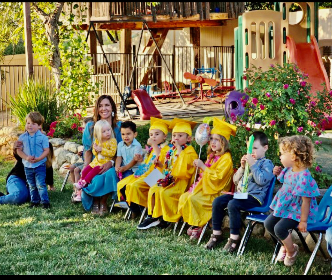 Miss Robin with preschool graduates in yellow caps and gowns in the sunny backyard play area